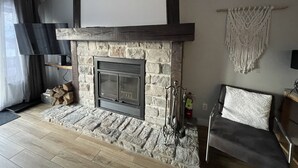 A living room with a stone fireplace, a flat-screen TV, a sofa, and a macrame wall hanging. - Ski in/ Ski out Modern 1 bedroom with fireplace (Mont-Tremblant)