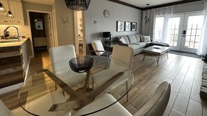 A modern living room with a glass dining table, a sofa, and a large window with curtains. - Ski in/ Ski out Modern 1 bedroom with fireplace (Mont-Tremblant)