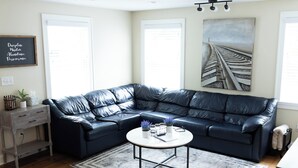 A modern living room with a black leather sectional sofa, a white coffee table, and a painting of a railway track.