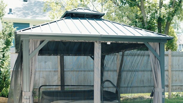 A covered outdoor seating area with a bench and a table, surrounded by a metal fence and a tree.