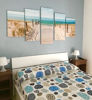 Quarto com cama, mesa de cabeceira, luminária e uma parede decorada com arte sobre o tema praia.