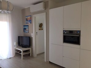 Uma sala de estar moderna com uma televisão de tela plana sobre um suporte branco, um forno embutido e uma porta que dá para outro cômodo.
