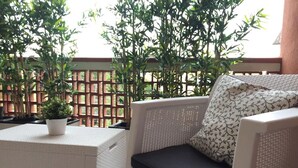 A wicker sofa with a black cushion and a patterned pillow, a small white table with a potted plant, and a wooden lattice fence in the background. - Bright, renovated apartment with two bedrooms and parking space - 300m from the sea (Grado)