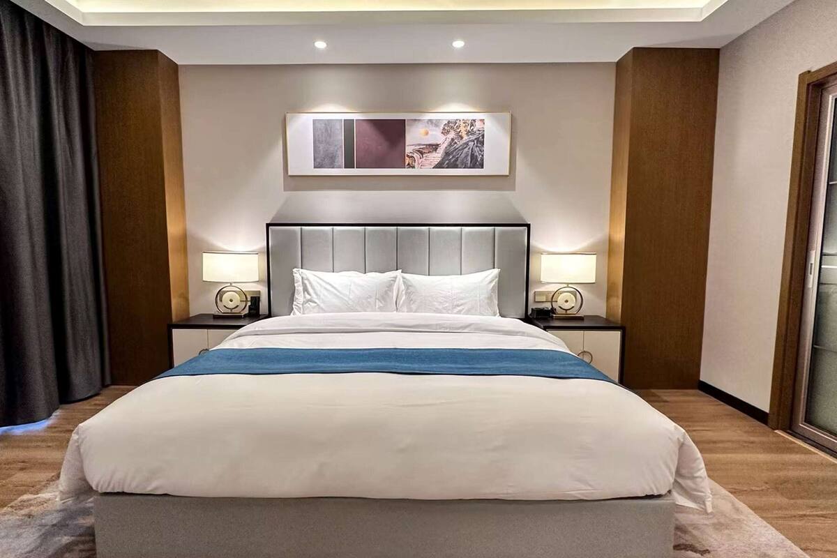 Une chambre d’hôtel avec un grand lit, deux tables de chevet avec des lampes, une œuvre d’art accrochée au mur et une fenêtre donnant sur un beau paysage.