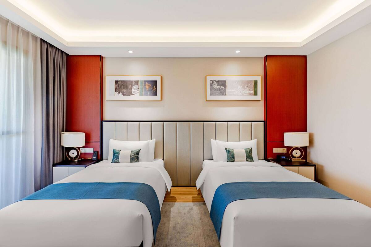 Une chambre d’hôtel avec deux lits, une tête de lit en bois, une grande fenêtre avec des rideaux et deux tableaux encadrés accrochés au mur.