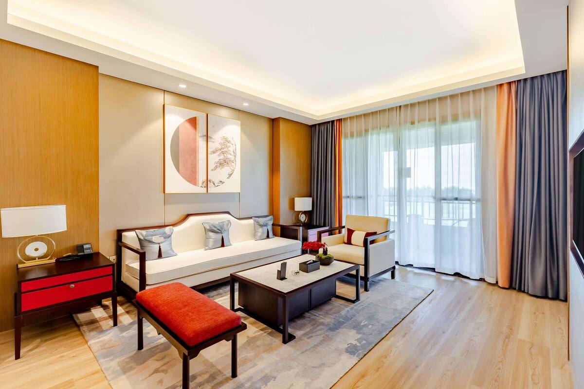Une chambre d’hôtel moderne avec un canapé, une table basse, un banc rouge, une télévision et une grande fenêtre avec des rideaux.