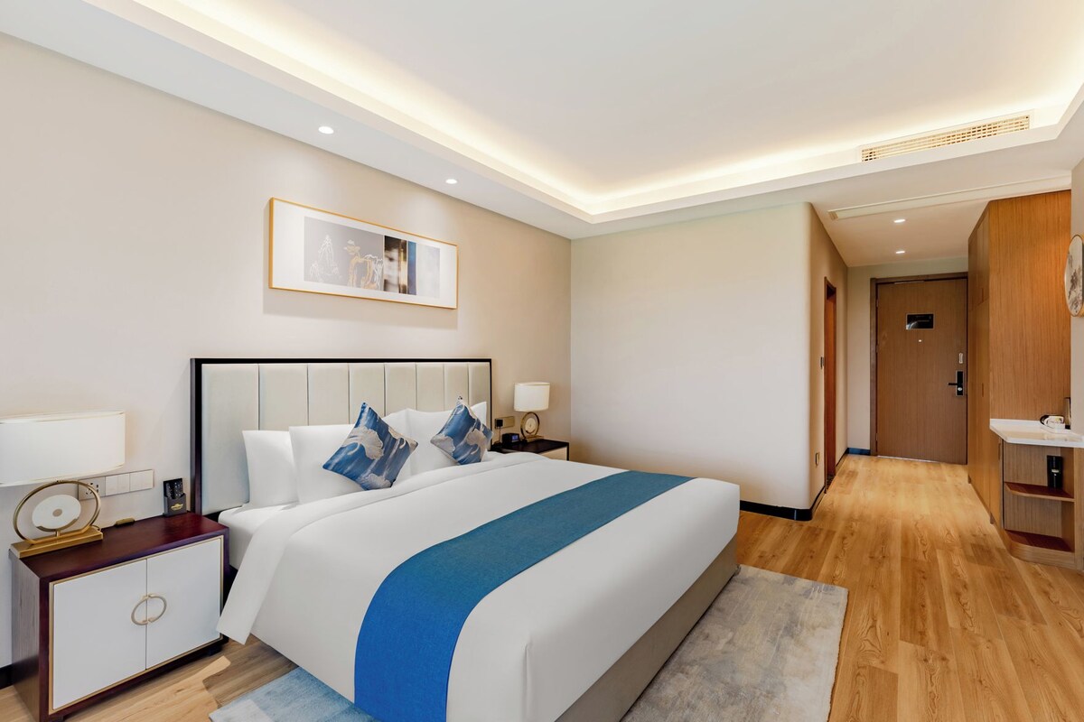A hotel room with a large bed, bedside tables, a nightstand, a desk, a chair, a mirror, a wardrobe, and a door.
