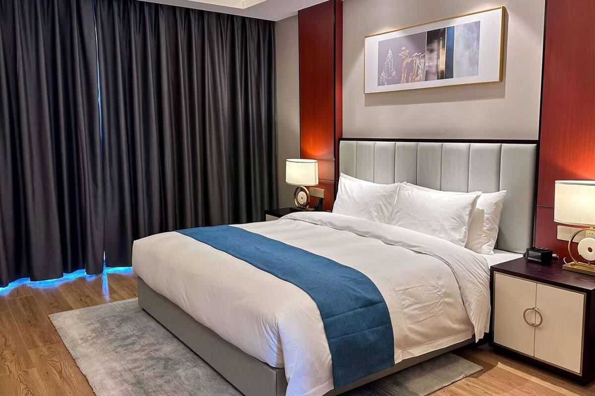 Une chambre d’hôtel avec un lit, une table de chevet, une lampe, un tableau au mur et une fenêtre avec des rideaux.