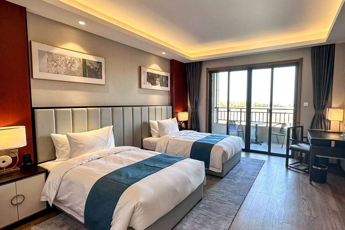 A hotel room with two beds, a desk, a chair, a balcony, and artwork on the walls.