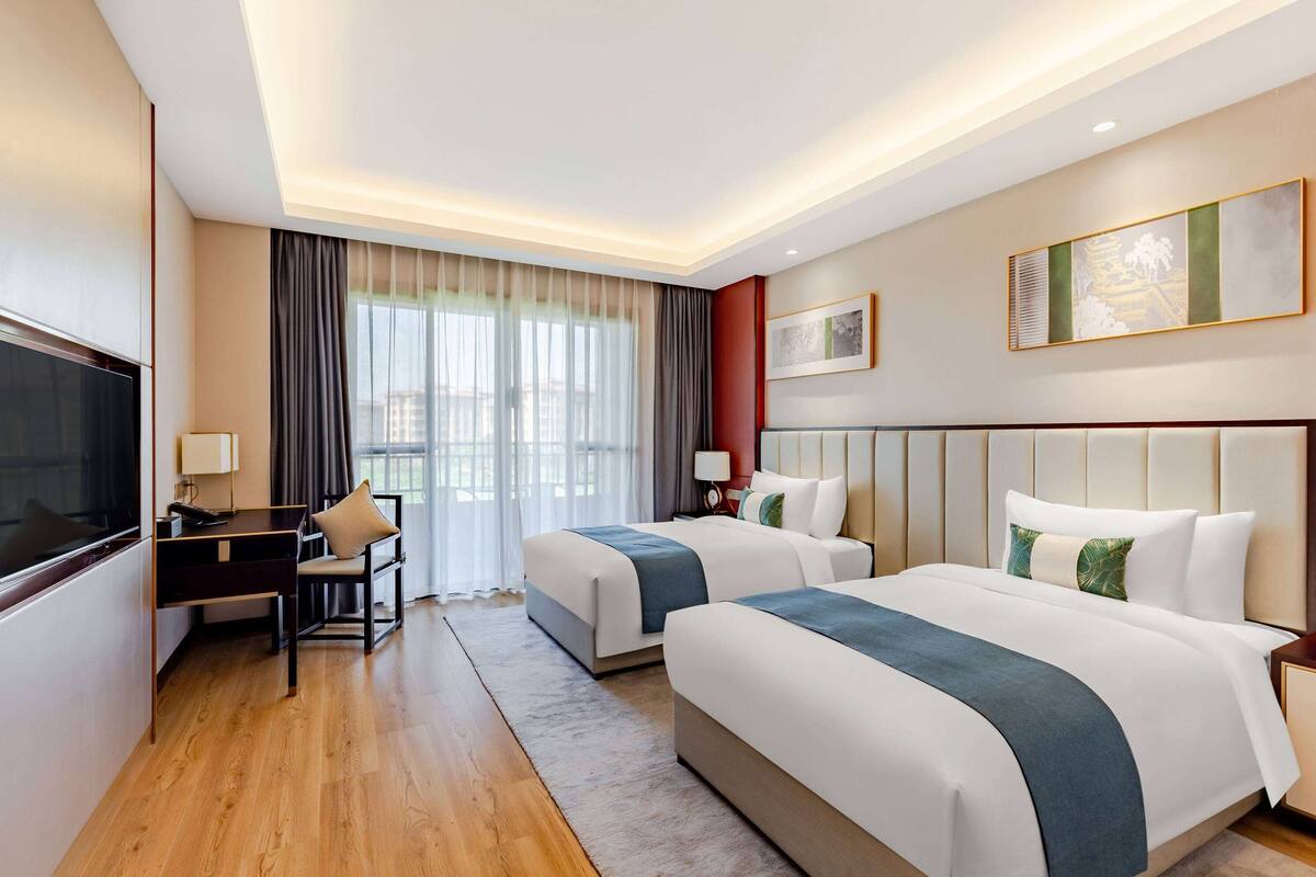 Une chambre d’hôtel avec deux lits, un bureau, une télévision et une grande fenêtre avec des rideaux.
