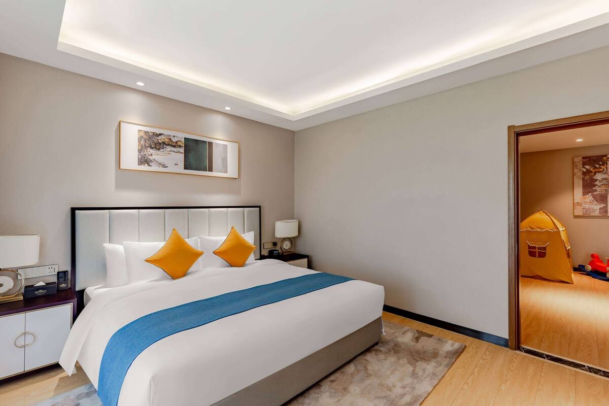 Une chambre d’hôtel moderne, dotée d’un grand lit, d’une table de chevet, d’une commode et d’une porte donnant sur une autre pièce.