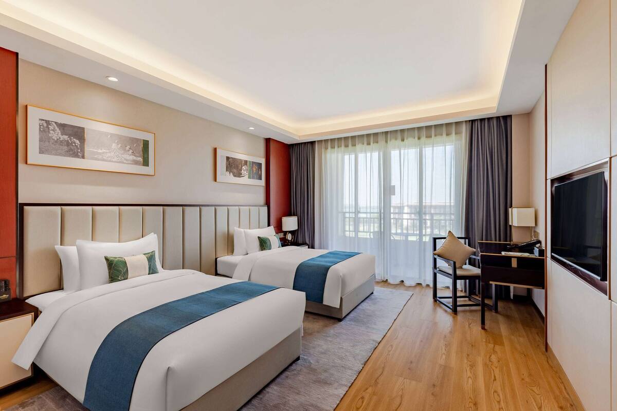 Une chambre d’hôtel avec deux lits, un bureau, une télévision et une grande fenêtre avec des rideaux.