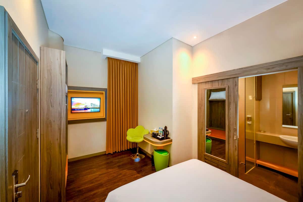 Quarto de hotel com guarda-roupa de madeira, pequena escrivaninha com cadeira, televisão na parede e banheiro com espelho e pia.