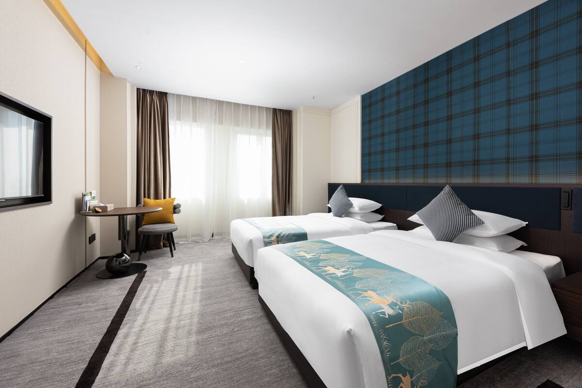 Ett hotellrum med två sängar, ett skrivbord, en stol och ett stort fönster med gardiner.