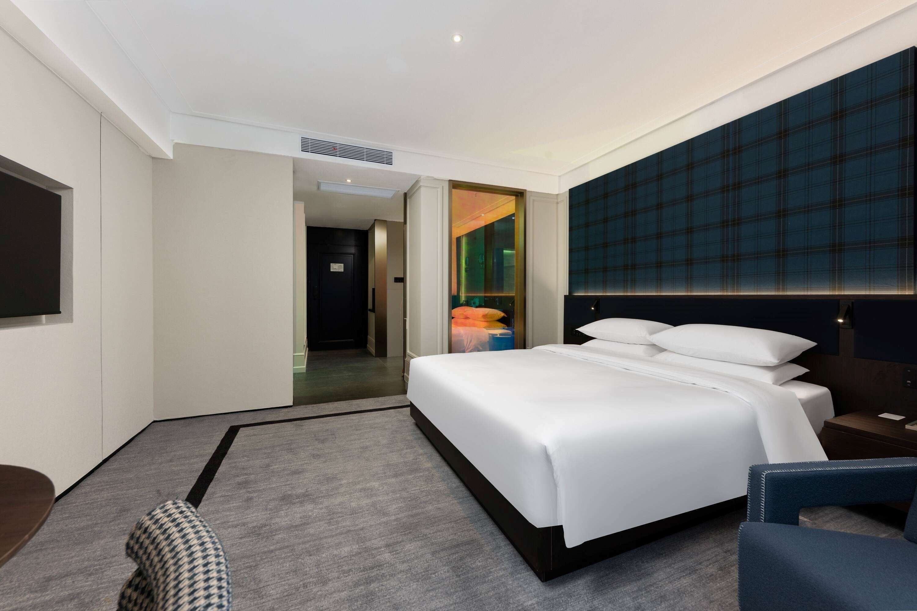 Habitación de hotel moderna con una cama grande, un escritorio y una silla. Se observa una vista del pasillo y otra habitación al fondo.