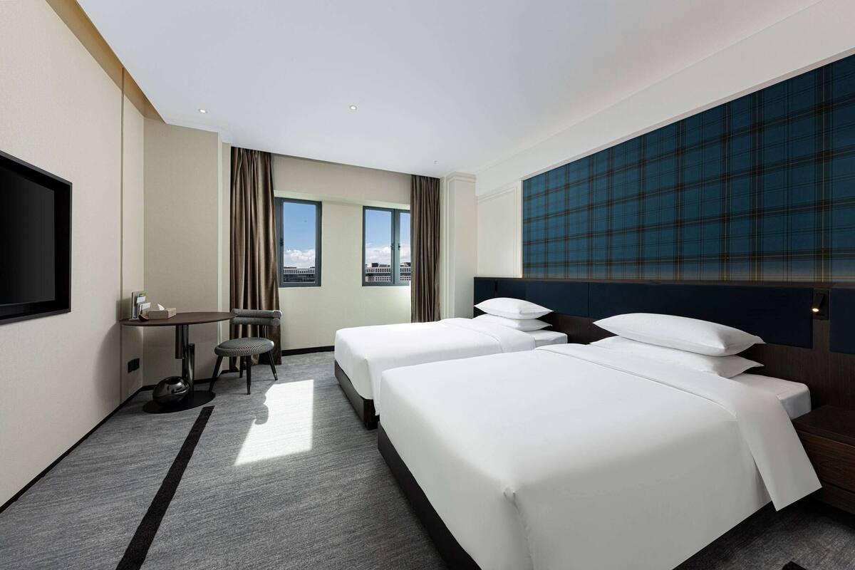 Ett hotellrum med två sängar, ett skrivbord, en stol, en TV och ett stort fönster med utsikt över staden.