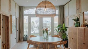 Un comedor con una mesa redonda, cuatro sillas, una lámpara colgante y vistas al exterior a través de ventanas amplias.