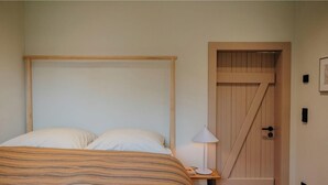 Un dormitorio con cabecera de cama de madera, dos almohadas, una mesita de noche con lámpara y una puerta de madera con ventanal.