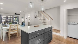 A modern kitchen with a central island, a dining area with a table and chairs, and a staircase leading to another level. - Modern Coastal 2BR: Walk to Beach, Pool at Back Door (Destin)