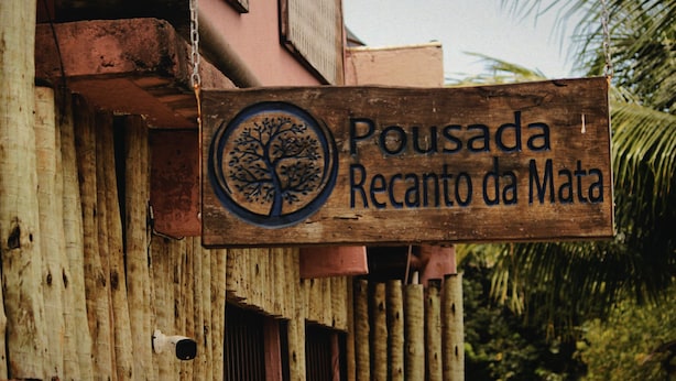 A wooden sign with a tree logo and text reading "Pousada Recanto da Mata". - Pousada Recanto da Mata (Porto Seguro)