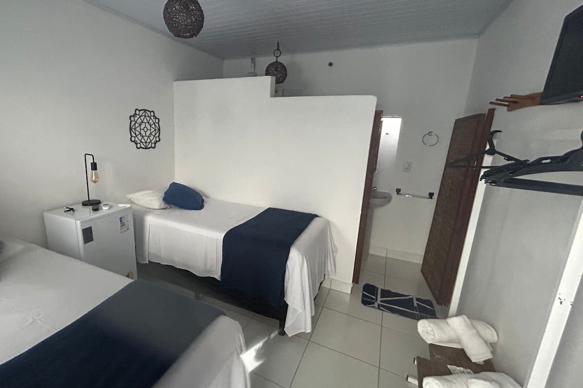 Quarto de hotel com duas camas, uma pequena mesa de cabeceira com abajur, um guarda-roupa e um banheiro visível através de uma porta aberta.