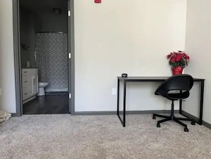 Quarto com escrivaninha, cadeira e planta em vaso. Banheiro visível por uma porta aberta.