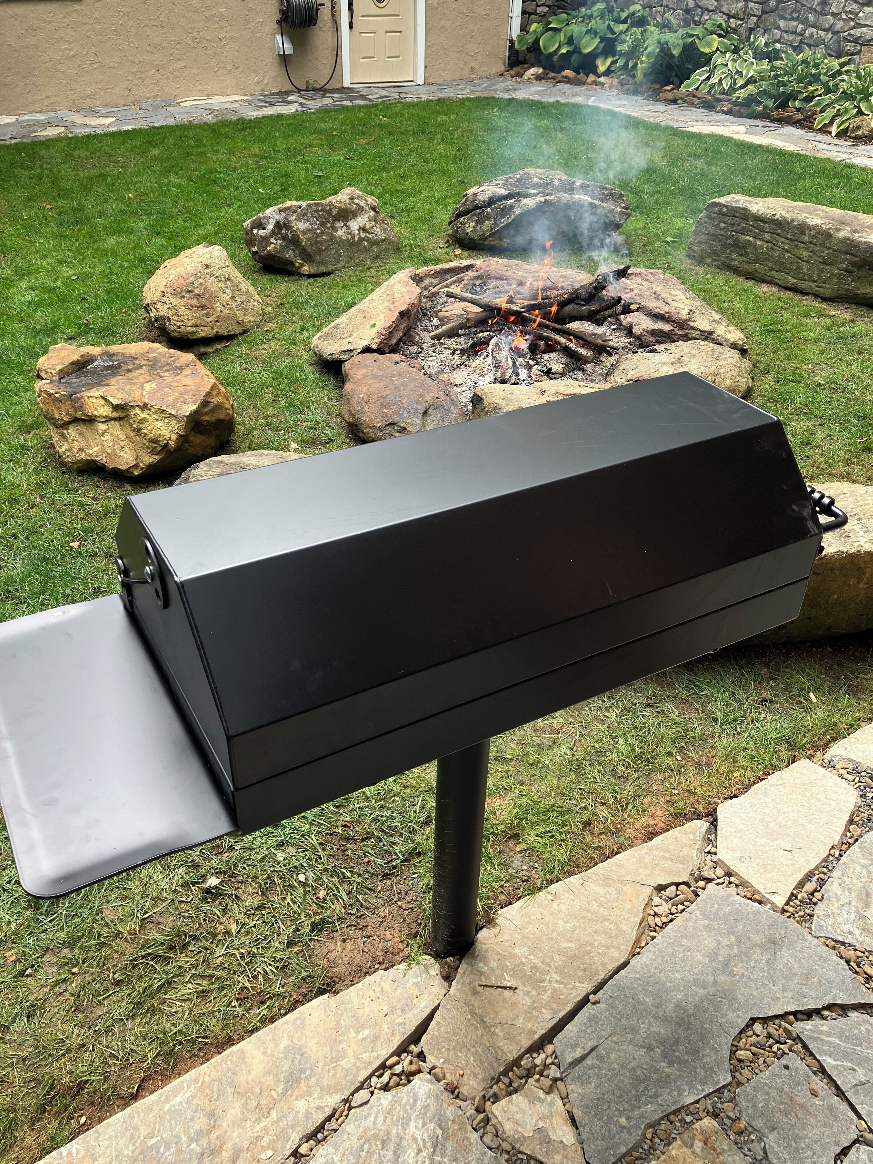 A black outdoor grill with a side burner, set on a stone patio with a grassy lawn and a stone wall in the background.