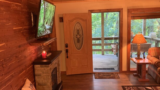 A cozy cabin interior with a stone fireplace, wooden walls, a television, a wooden door leading to a deck, and a plaid sofa. - BEARADISE 1 - Five Min Walk To Downtown Banner Elk (Banner Elk)