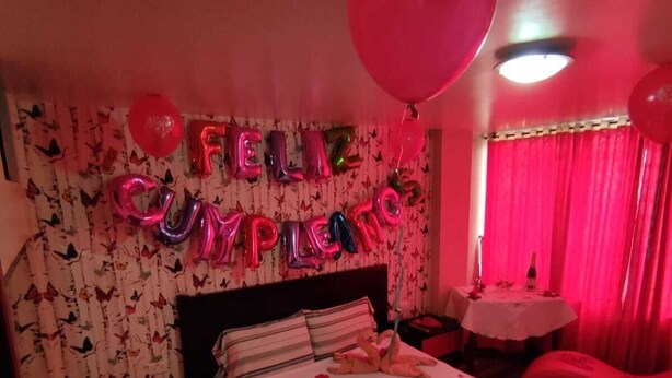 Una habitación con una cama, una pared con globos que deletrean "FELIZ CUMPLEAÑOS" y un mensaje sobre la cama.