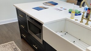 Eine Küche mit weißer Arbeitsplatte, schwarzen Schränken, einer Edelstahl-Spüle und einem gemusterten Teppich.