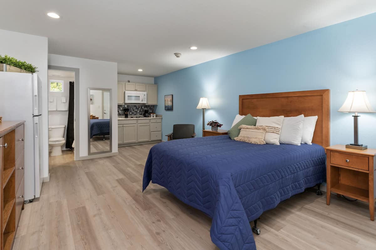 A bedroom with a large bed, wooden headboard, bedside tables with lamps, and a view into the adjacent kitchen.