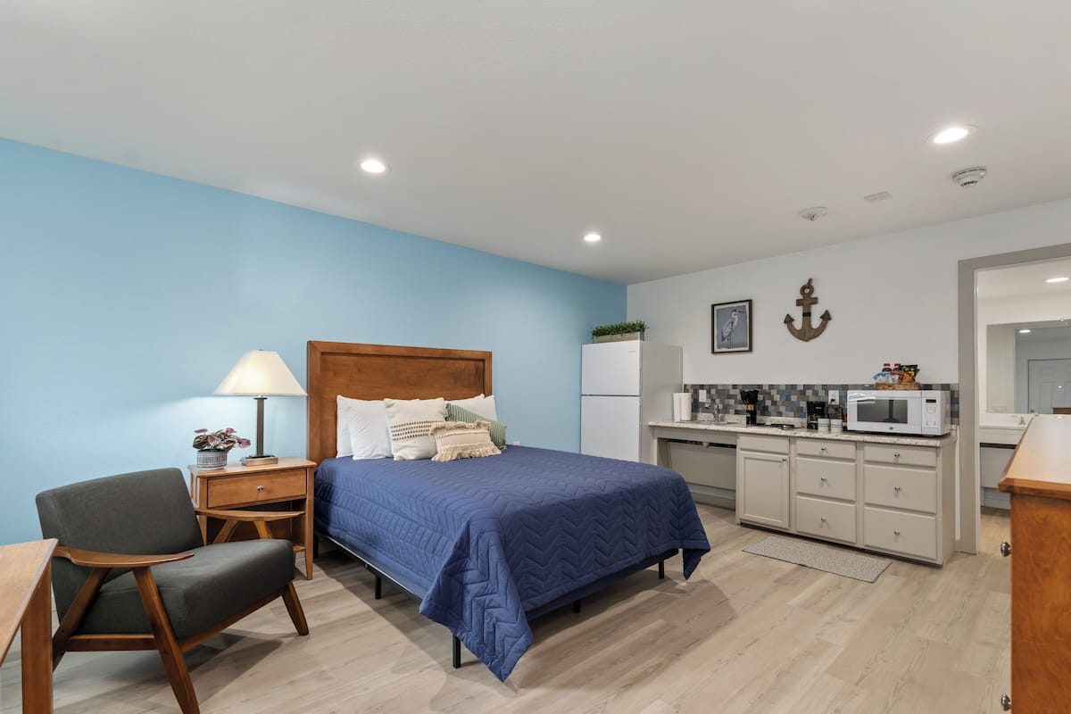 A bedroom with a bed, bedside table, lamp, chair, and a kitchenette area with a microwave.