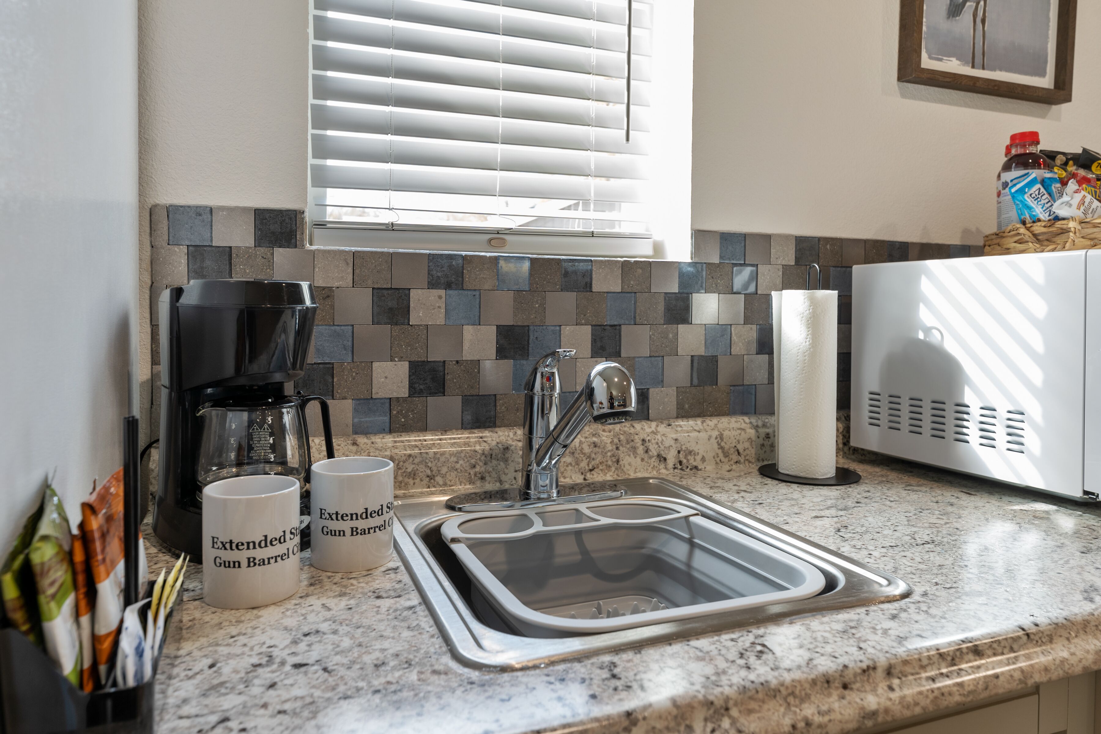 A kitchen with a granite countertop, a stainless steel sink, a coffee maker, and two mugs labeled "Extended Gun Barrel."