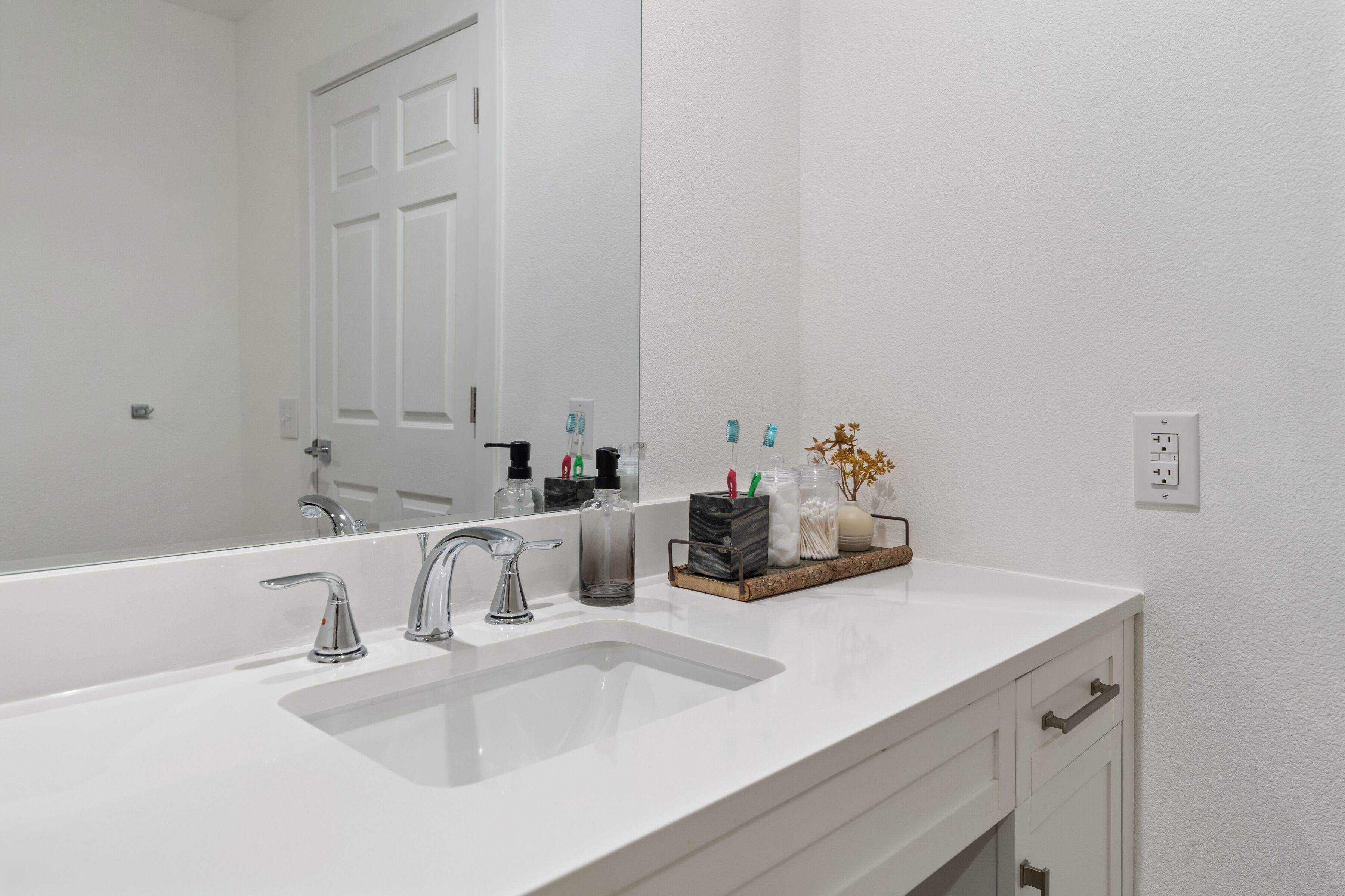 Un baño moderno con un lavamanos grande de color blanco, dos llaves, un espejo y una encimera con artículos de aseo.
