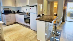 A modern kitchen with white cabinets, a granite countertop, and stainless steel appliances. There are three bar stools near the counter. - Central Two Bedroom Apartment on the Second Line of the Beach in Marbella. (Marbella)
