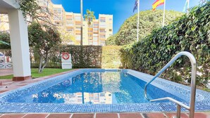 A swimming pool with a tiled deck, surrounded by greenery and a building in the background. - Central Two Bedroom Apartment on the Second Line of the Beach in Marbella. (Marbella)