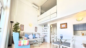 Een moderne woonkamer met een bank, een salontafel en een eethoek met stoelen. Er is een plant, een boekenkast en een zichtbare keuken.