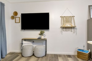 Una habitación con una televisión de pantalla plana montada en la pared, una pequeña consola de madera con una planta, dos otomanas grises, un tapiz de macramé y una cesta de mimbre en el suelo.
