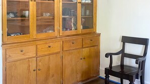Un armari de fusta amb portes de vidre que conté vaixelles de cuina, una cadira negra i un llum penjant al sostre.