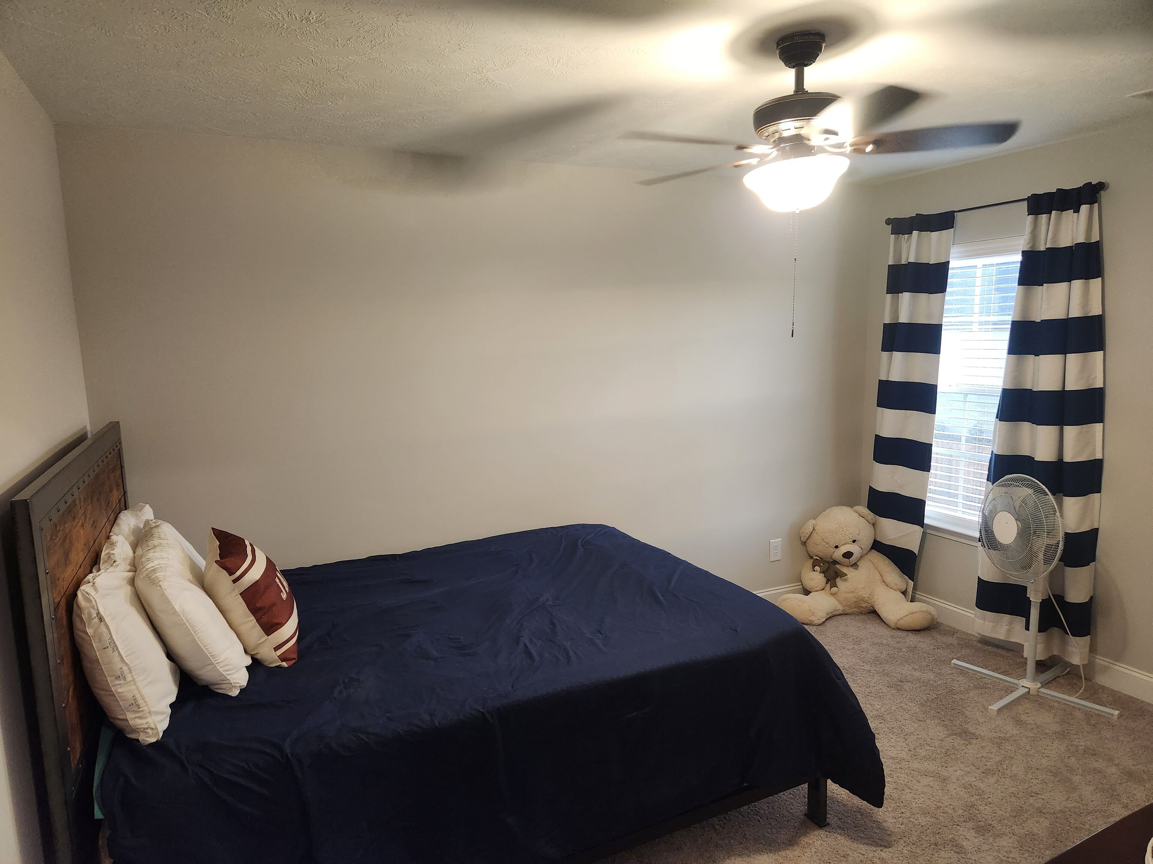 Ein Schlafzimmer mit Bett, Kissen, Deckenventilator, Fenster mit Vorhängen und einem Teddybär.
