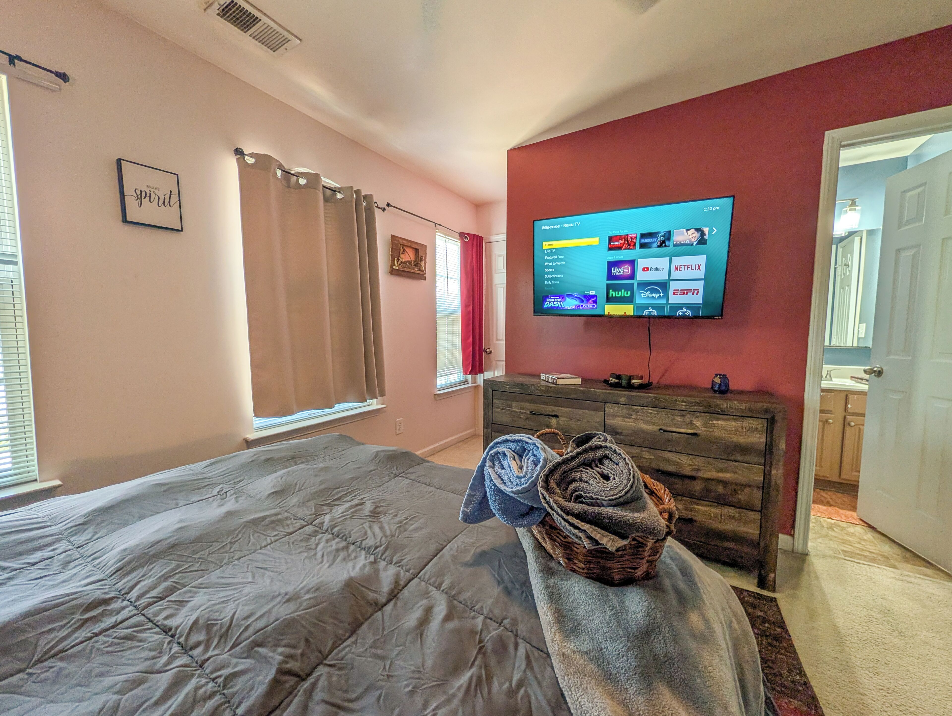 A bedroom with a bed, a TV mounted on the wall, a dresser, and a window with curtains.