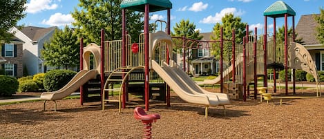 A playground with slides and climbing structures, surrounded by a grassy area and trees.