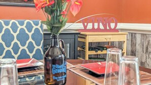 Un comedor con una mesa dispuesta para dos, un florero con flores rojas, un letrero de madera con la palabra 'VINO' y una botella etiquetada 'The Barrel Grouse'.
