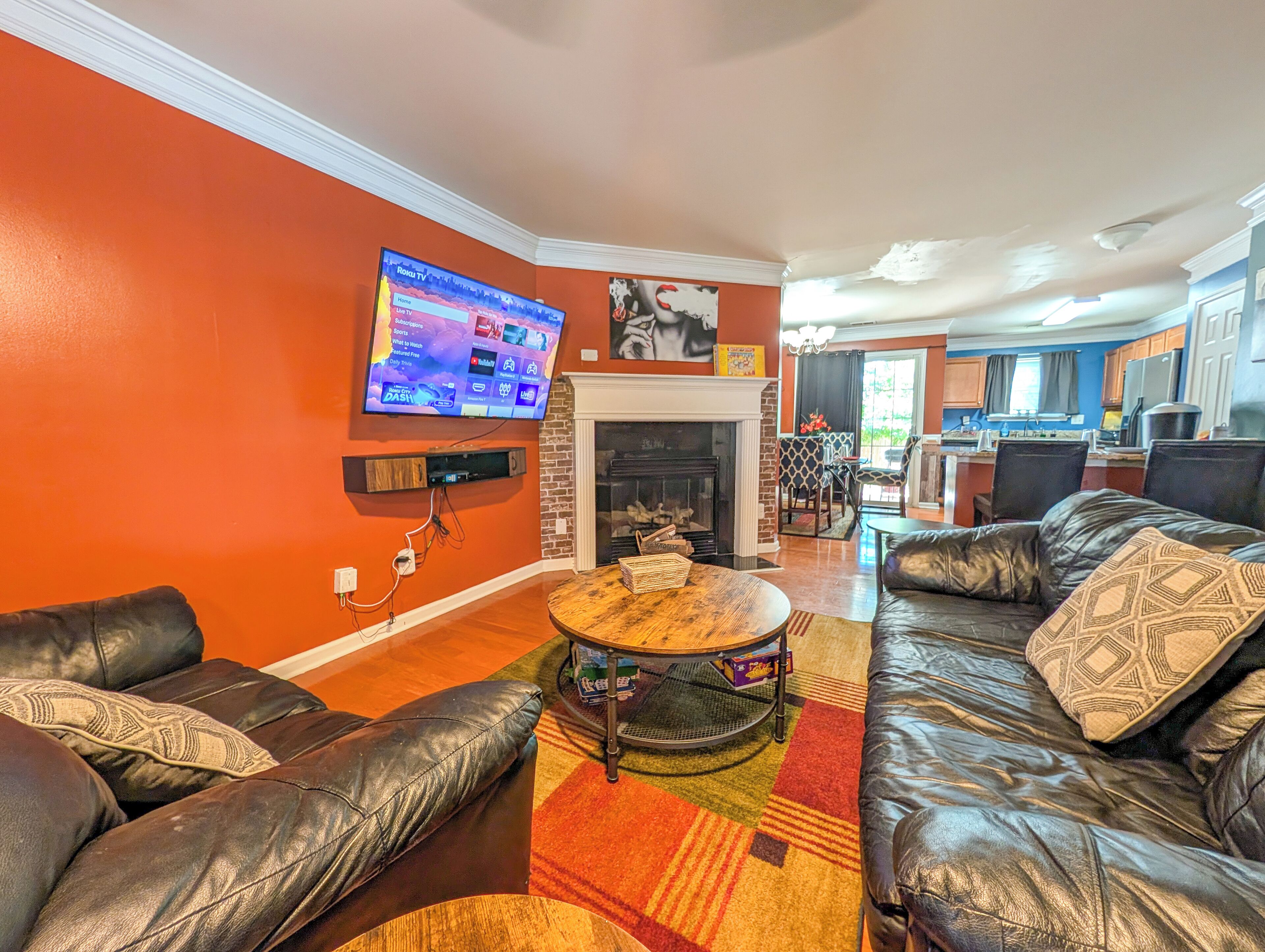 A living room with a fireplace, leather sofas, a television mounted on the wall, and a kitchen in the background.