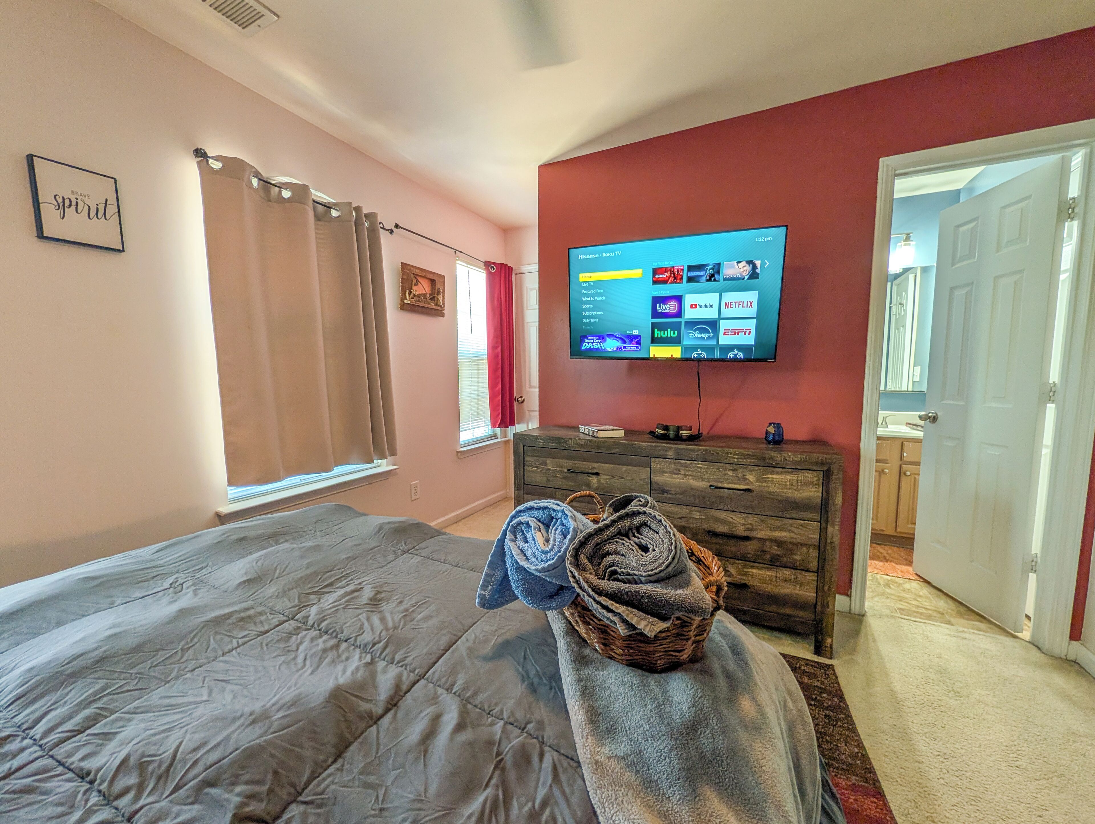 A bedroom with a bed, a TV mounted on the wall, a dresser, and a window with curtains.