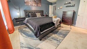 Un dormitorio con cama, mesitas de noche, cómoda, ventilador de techo y una obra de arte con un motivo urbano en la pared.