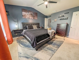 Une chambre à coucher comprenant un lit, des tables de chevet, une commode, un ventilateur de plafond et une œuvre d’art représentant un paysage urbain accrochée au mur.