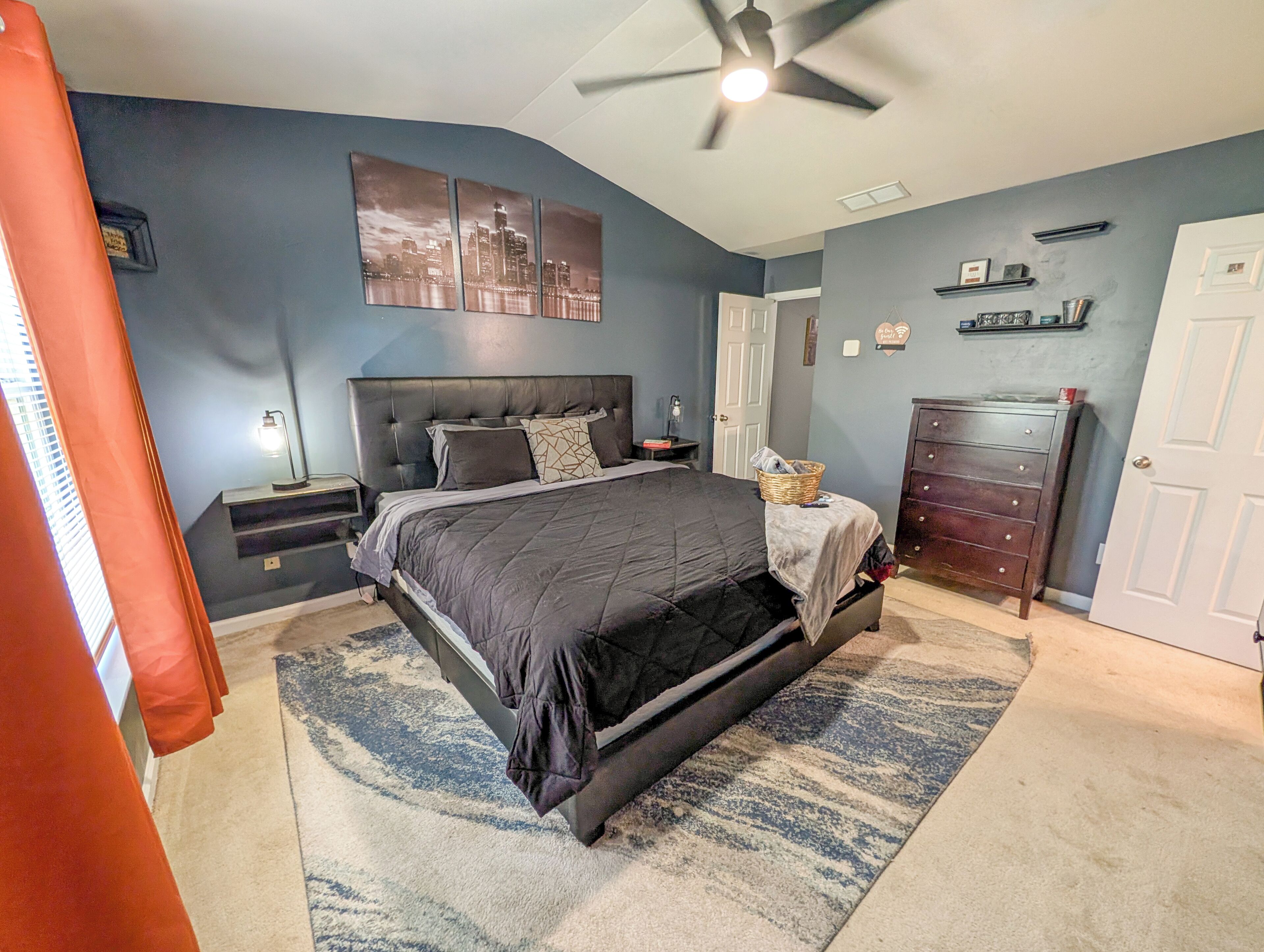 A bedroom with a bed, nightstands, a dresser, a ceiling fan, and a cityscape artwork on the wall.