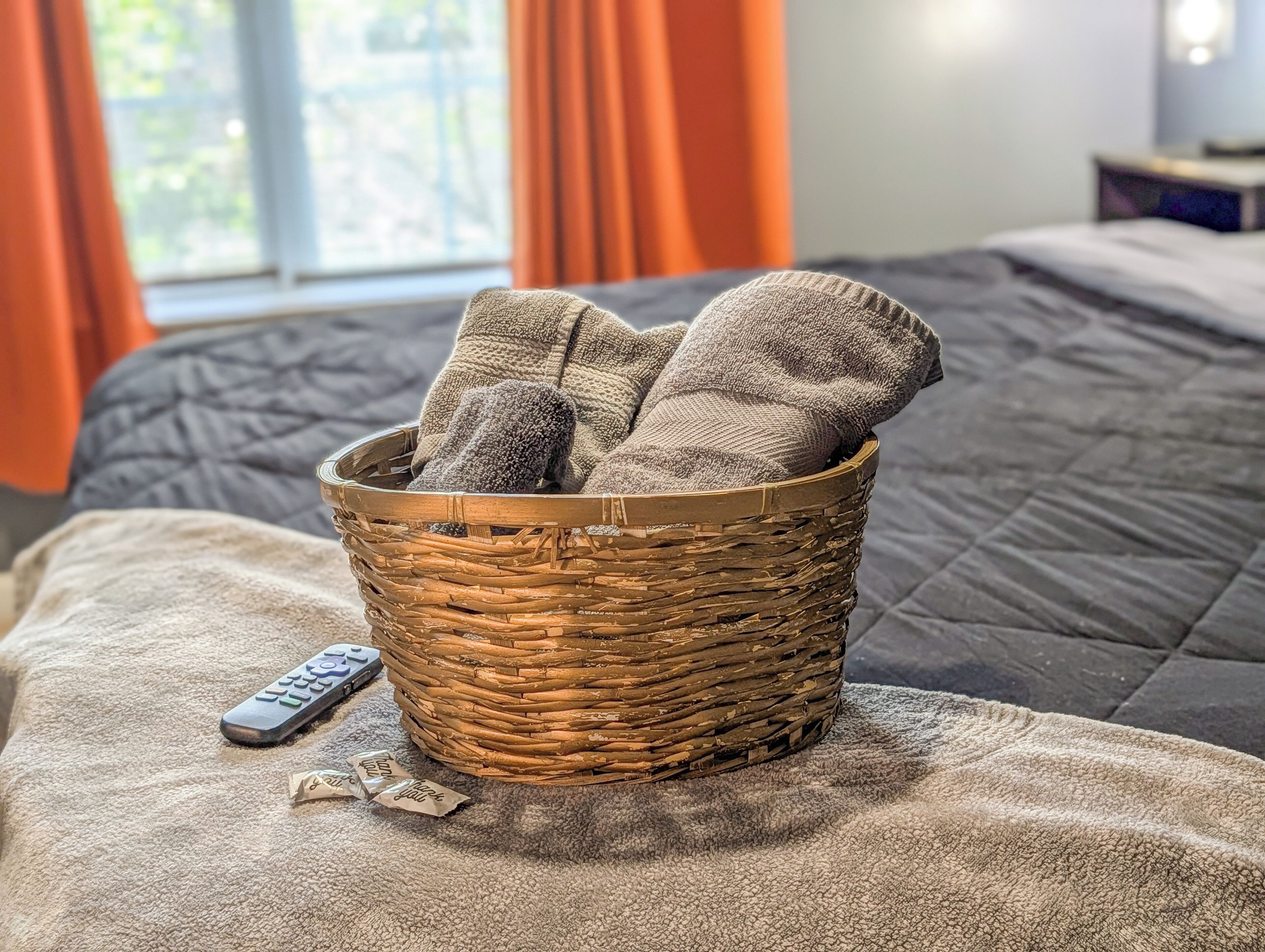 Un panier en osier rempli de chaussettes est posé sur un lit, à côté duquel se trouvent une télécommande et une boîte de mouchoirs.