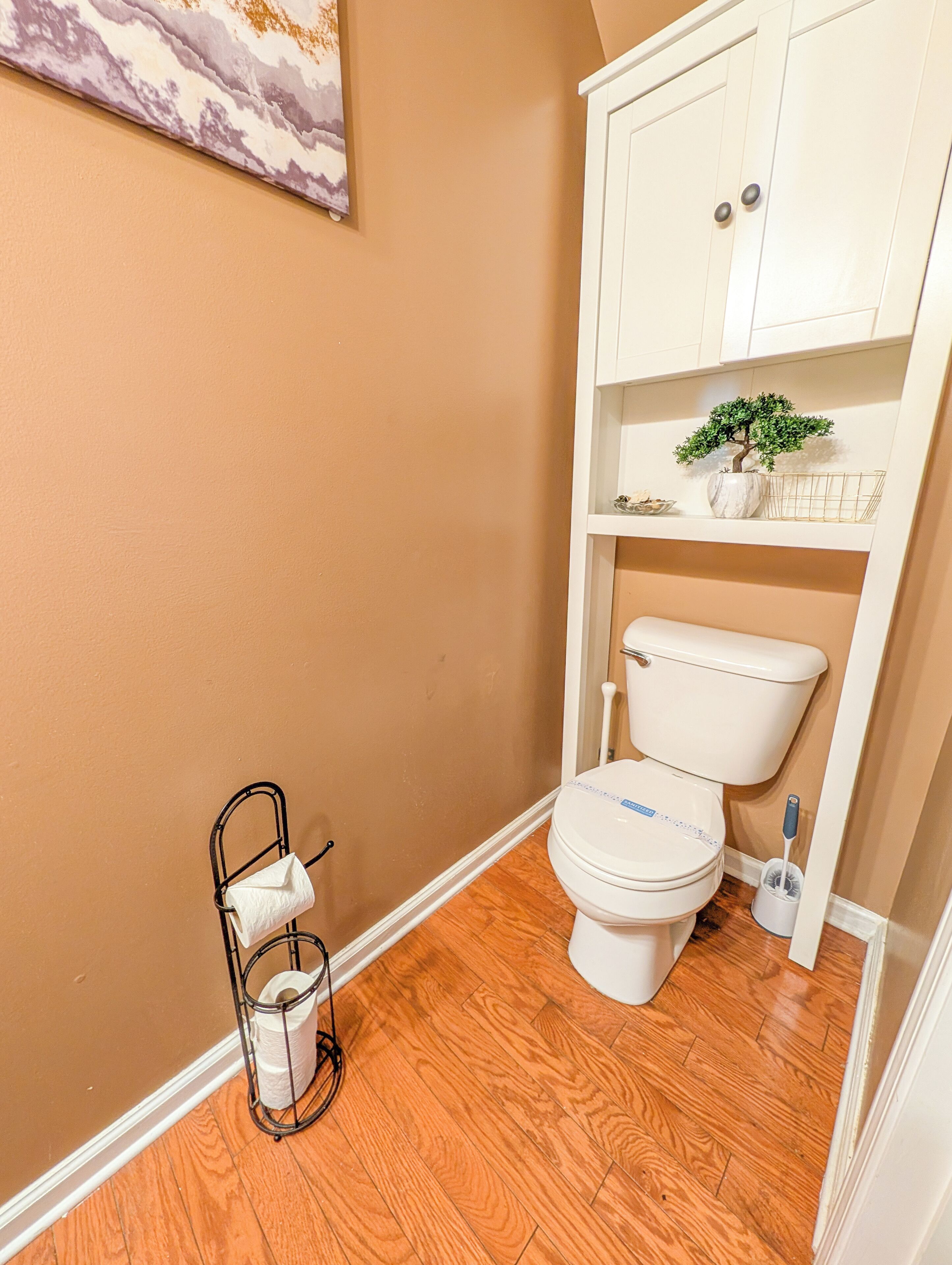 Une salle de bain avec des toilettes, un porte-mouchoirs en métal, une décoration murale à motifs de marbre, un meuble avec une plante et un plancher en bois.